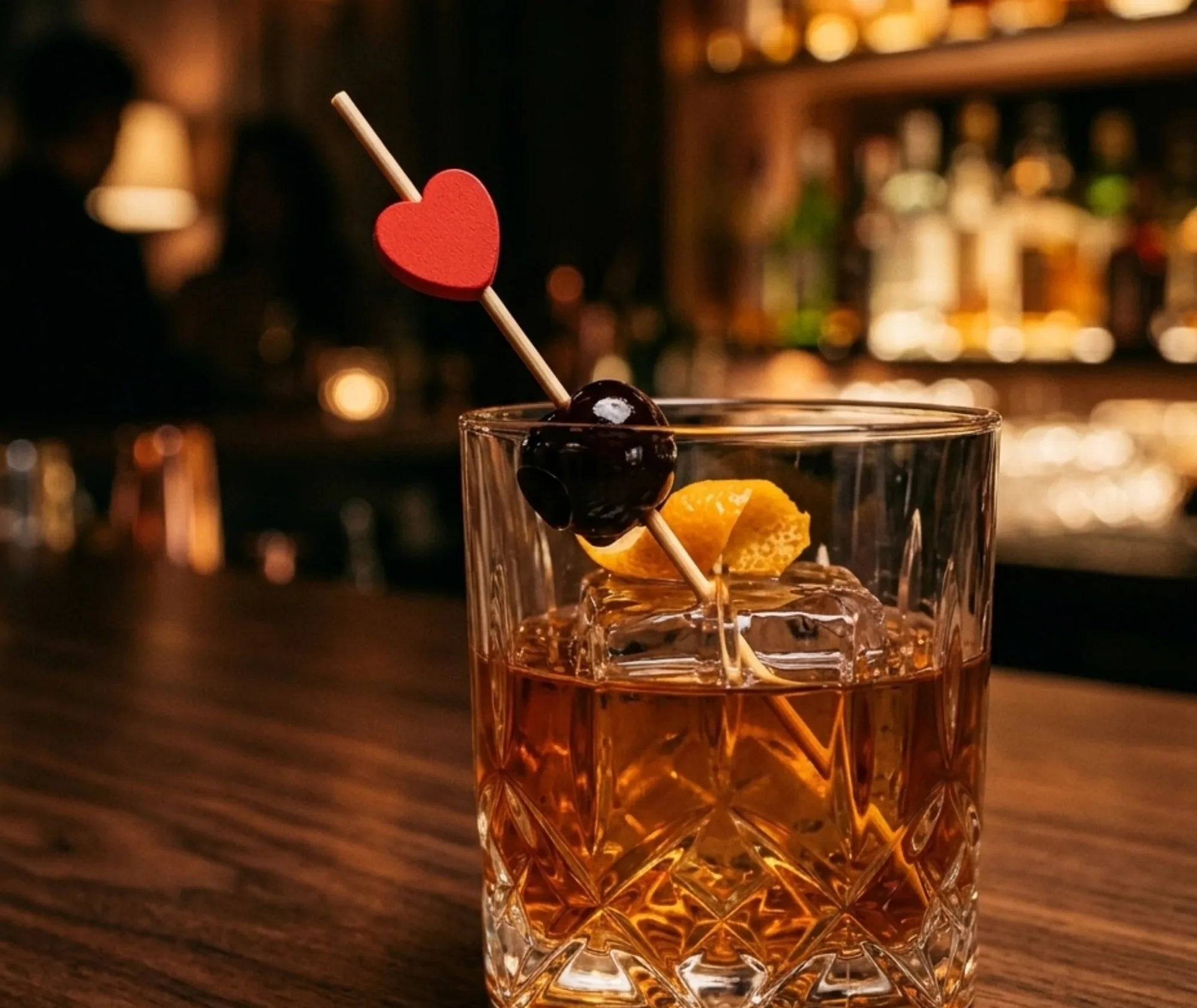 Pique à cocktail cœur rouge plantée dans un Old Fashioned garni d'une cerise et d'un zeste d'orange, au comptoir d'un bar