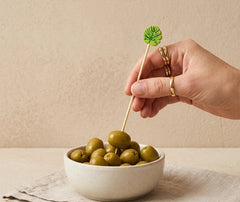 Main portant des bagues dorées piquant une olive verte avec un pique à cocktail feuille, au-dessus d'un bol d'olives sur fond beige.