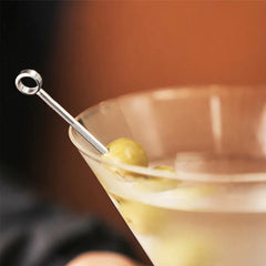 Gros plan macro d'une olive verte piquée sur une pique argentée, posée sur le bord d'un verre à martini.