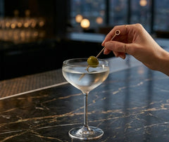Main féminine plaçant une olive sur un pique doré dans un verre à martini, dans un bar sombre et élégant.