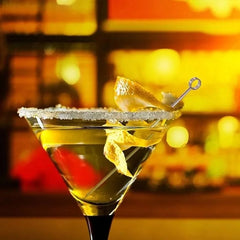 Verre à martini avec zeste de citron piqué sur une pique argentée, posé sur un bar flou en arrière-plan doré.