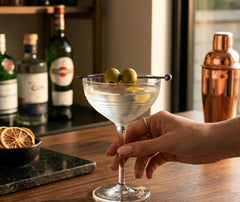 Pic à cocktail irisé piquant deux olives vertes dans un verre à martini tenu à la main, avec des bouteilles de bar en arrière-plan.