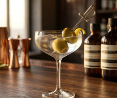 Pic à cocktail épée piqué dans des olives vertes et un zeste de citron sur un verre à Martini, avec des jiggers en cuivre en arrière-plan.