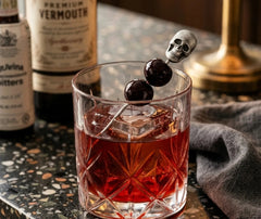 Deux pics à cocktail tête de mort piqués dans des cerises au-dessus d'un Negroni, avec une bouteille de vermouth en arrière-plan.