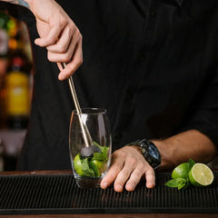 Un barman en chemise noire prépare un cocktail au comptoir en écrasant de la menthe et du citron vert avec un pilon doré.