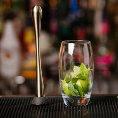 Pilon à cocktail argenté posé debout à côté d'un verre garni de menthe et de citron vert, sur un comptoir de bar à l'ambiance tamisée.