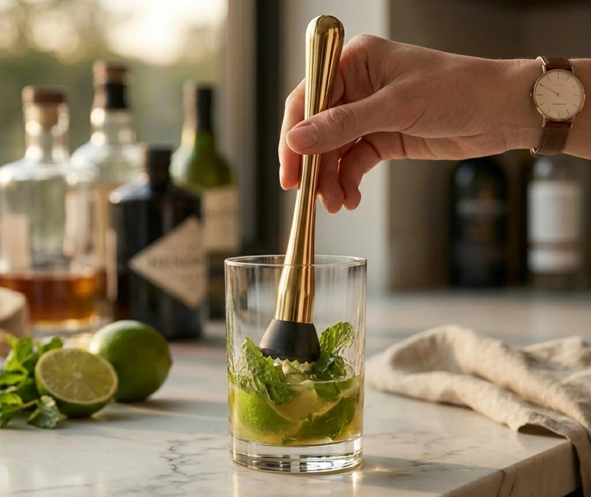 Une main portant une montre tient un pilon doré au-dessus d'un verre garni de feuilles de menthe et de citron vert dans un bar ensoleillé.