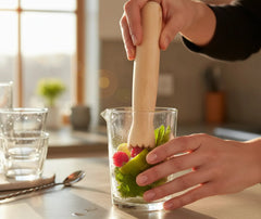Une main utilise le pilon en bois pour piler menthe, citron vert et framboises dans un verre à mélange.