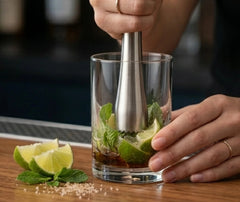 Une main pile menthe et citron vert avec le pilon dans un verre de mojito, sur un comptoir en bois avec quartiers de citron vert et sucre.