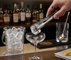 Pince à glace cocktail déposant un glaçon dans une coupe à cocktail, entourée de bouteilles de spiritueux