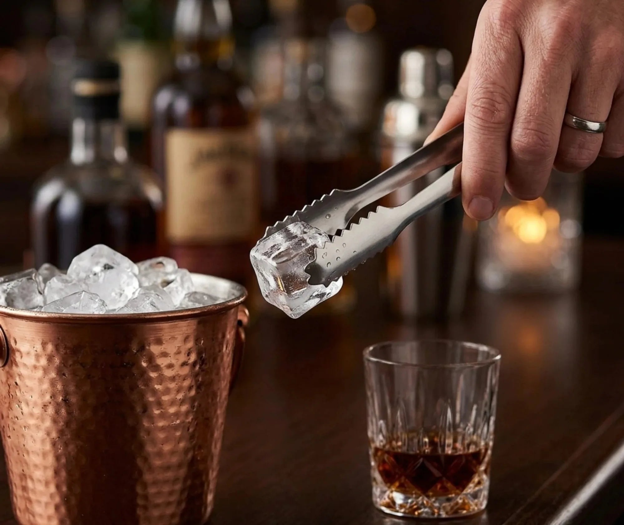 Pince à glace cocktail saisissant un glaçon dans un seau en cuivre, avec un verre de whisky sur le comptoir