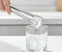Main utilisant la pince à glace inox pour ajouter un glaçon dans un verre d'eau, dans une cuisine lumineuse.