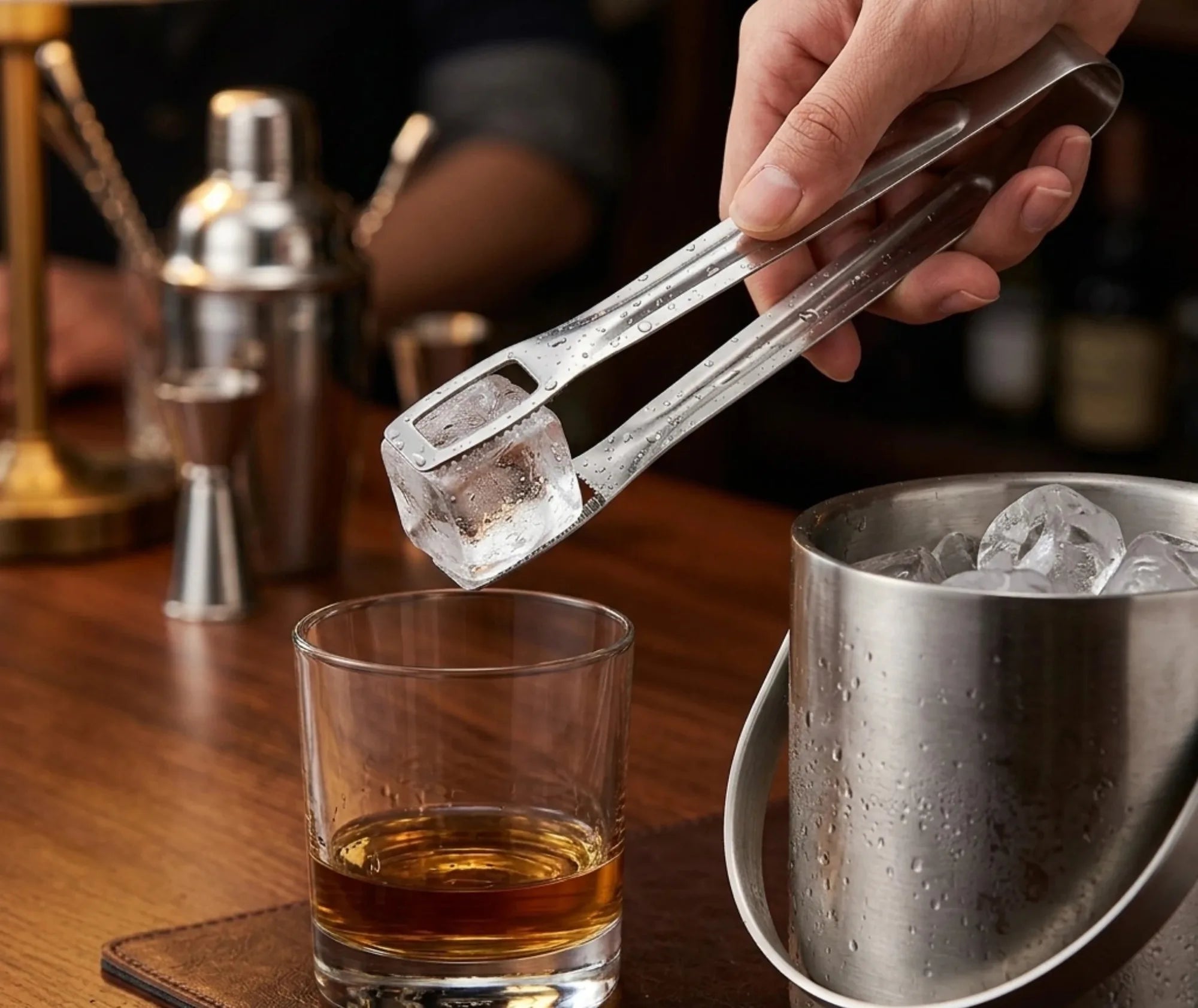 Main tenant la pince à glace inox pour déposer un glaçon dans un verre de whisky, ambiance bar en bois avec shaker en arrière-plan.