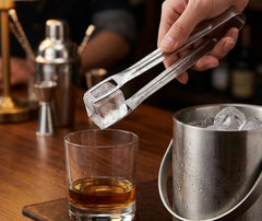 Main tenant la pince à glace inox pour déposer un glaçon dans un verre de whisky, ambiance bar en bois avec shaker en arrière-plan.