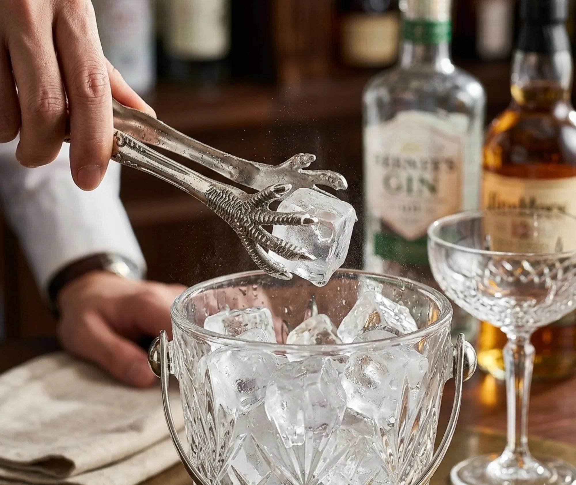 Main tenant la pince serres d'aigle et déposant un glaçon dans un seau, bouteilles de gin en arrière-plan de bar
