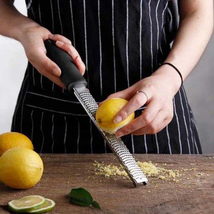 Personne en tablier rayé râpant le zeste d'un citron jaune sur une planche en bois, entourée de citrons et d'un citron vert coupé