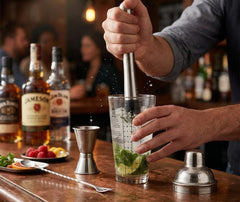 Utilisation du pilon du set barman cocktail pour piler la menthe et le citron vert sur un comptoir de bar en bois, avec doseur et fruits rouges