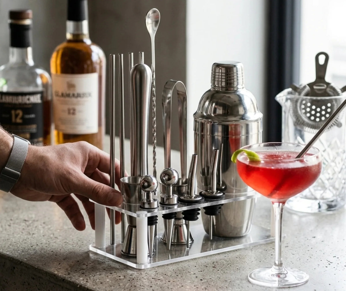 Barman attrapant un accessoire du kit cocktail posé devant des bouteilles de whisky, avec un cocktail rouge au premier plan.