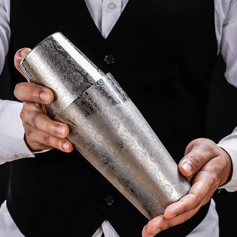 Un barman en gilet noir tient le shaker Boston De Luxe gravé entre ses deux mains pour préparer un cocktail.