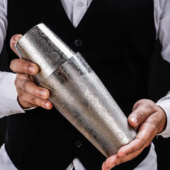 Un barman en gilet noir tient le shaker Boston De Luxe gravé entre ses deux mains pour préparer un cocktail.