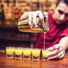 Barman utilisant le shaker Boston doré pour verser un cocktail dans une série de verres à shot