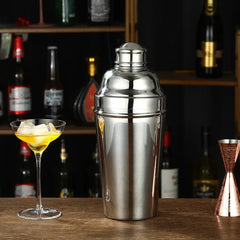 Shaker à cocktail en inox 1L posé sur un comptoir de bar en bois, à côté d'un verre à cocktail garni de glaçons et d'un doseur en cuivre, avec des bouteilles en arrière-plan.