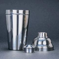 Shaker à cocktail en inox 1L démonté en trois parties : le grand gobelet, le couvercle filtre et le petit bouchon, sur fond gris uni.