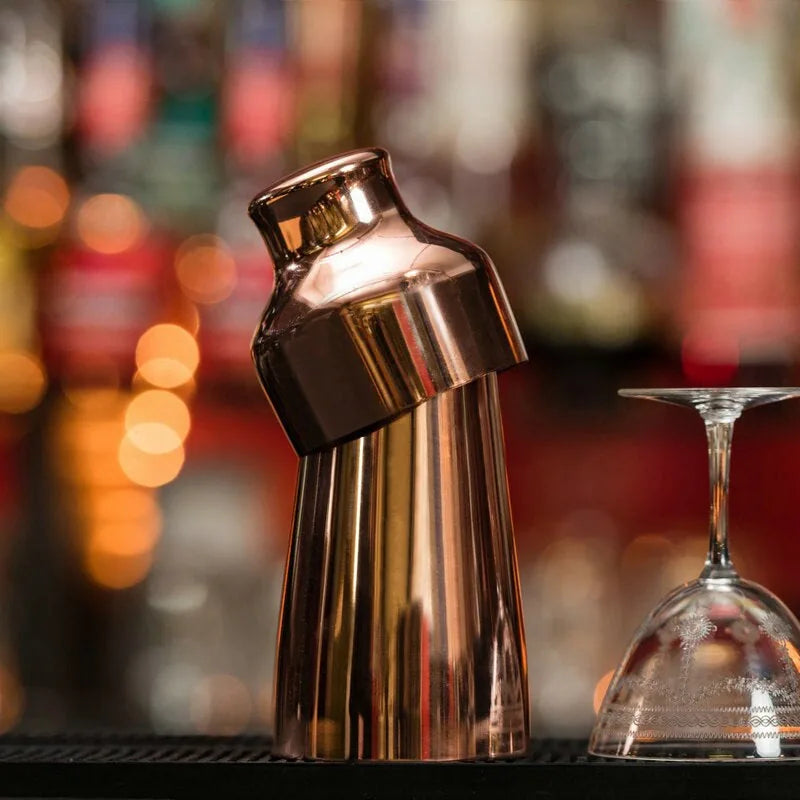 Shaker cocktail bar en cuivre incliné sur un bar, à côté de verres à cocktail retournés