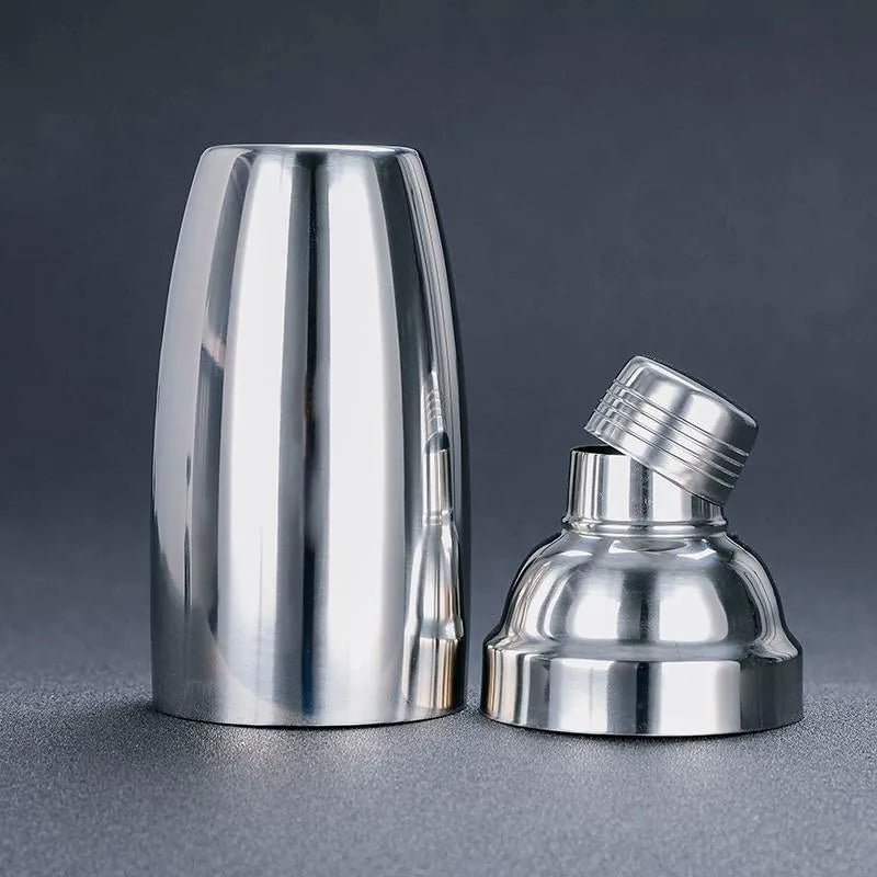 Shaker cocktail classique en inox avec le bouchon doseur dévissé et incliné sur la base dôme, à côté du gobelet cylindrique.