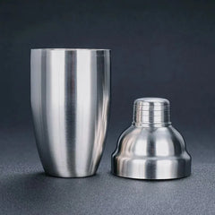 Shaker cocktail classique en inox démonté en deux parties : le gobelet cylindrique et la base dôme avec son bouchon vissé, sur fond sombre.