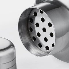 Gros plan sur la passoire perforée en inox du shaker cocktail classique, avec le bouchon doseur posé à côté.