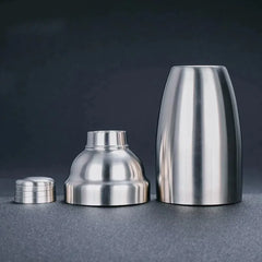 Les trois éléments du shaker cocktail classique en inox séparés : corps, passoire et bouchon doseur, sur fond sombre.