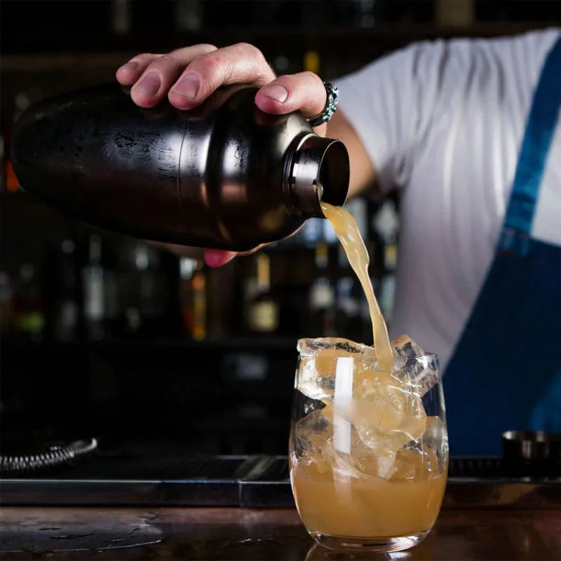 Barman versant un cocktail depuis le shaker noir dans un verre rempli de glace, en situation au bar.