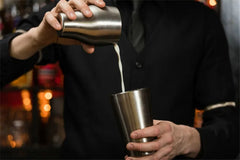 Barman en tenue noire versant un cocktail depuis le shaker argenté dans un verre, gros plan sur les mains