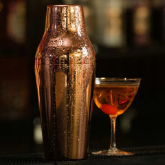 Shaker cocktail cuivré couvert de gouttes de condensation posé à côté d'un cocktail ambré