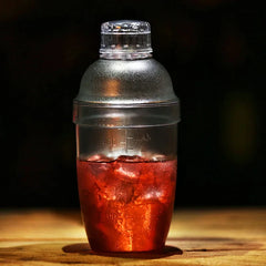 Shaker à cocktail gradué de 550 ml posé sur une table en bois, rempli d'un cocktail rouge glacé en ambiance tamisée.
