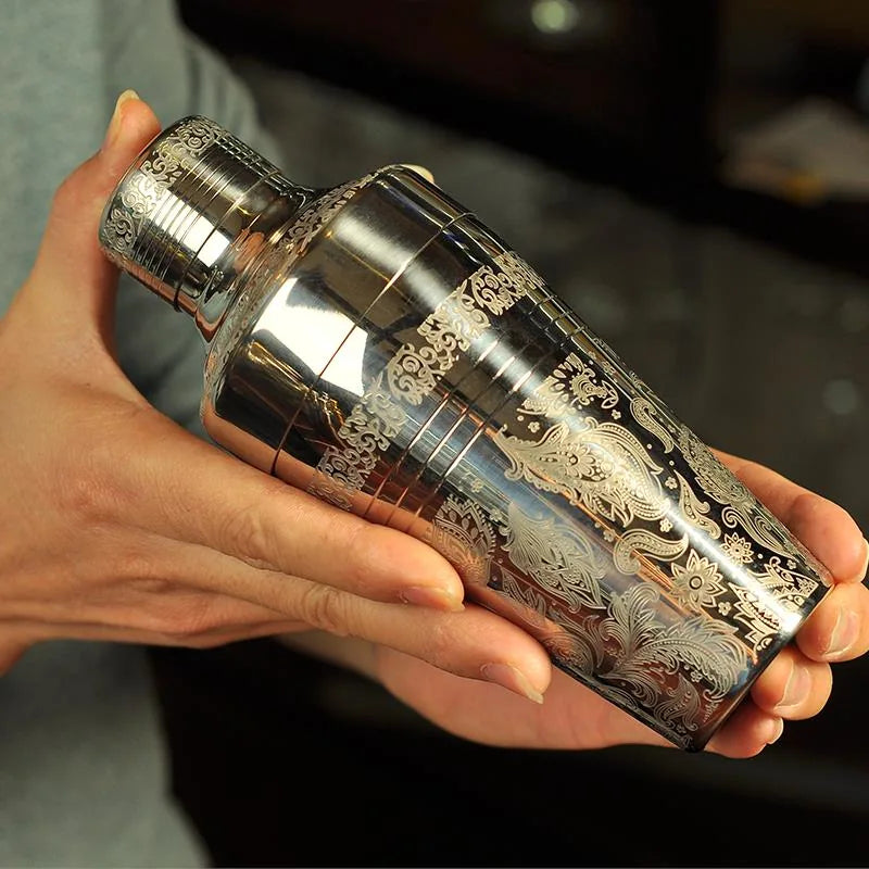 Gros plan du shaker cocktail gravé tenu en main, détail de la gravure florale