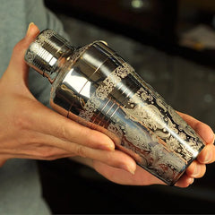 Gros plan du shaker cocktail gravé tenu en main, détail de la gravure florale