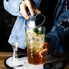 Une personne en veste en jean prépare un cocktail avec le shaker en plastique sur un plateau de bar, garni de zeste de citron et de menthe.