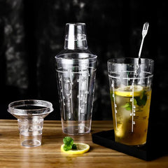 Set complet du shaker à cocktail en plastique avec sa mesurette et un verre à cocktail garni de citron et de menthe sur un comptoir en bois.
