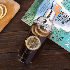 Shaker à cocktail rempli de thé glacé et de tranches de citron, posé sur une table en bois avec un magazine et une planche à découper en arrière-plan
