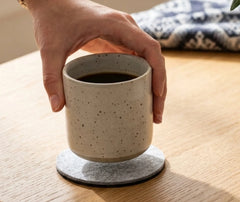 Main posant une tasse en céramique tachetée remplie de café sur un sous-verre en feutrine, sur une table en bois clair.