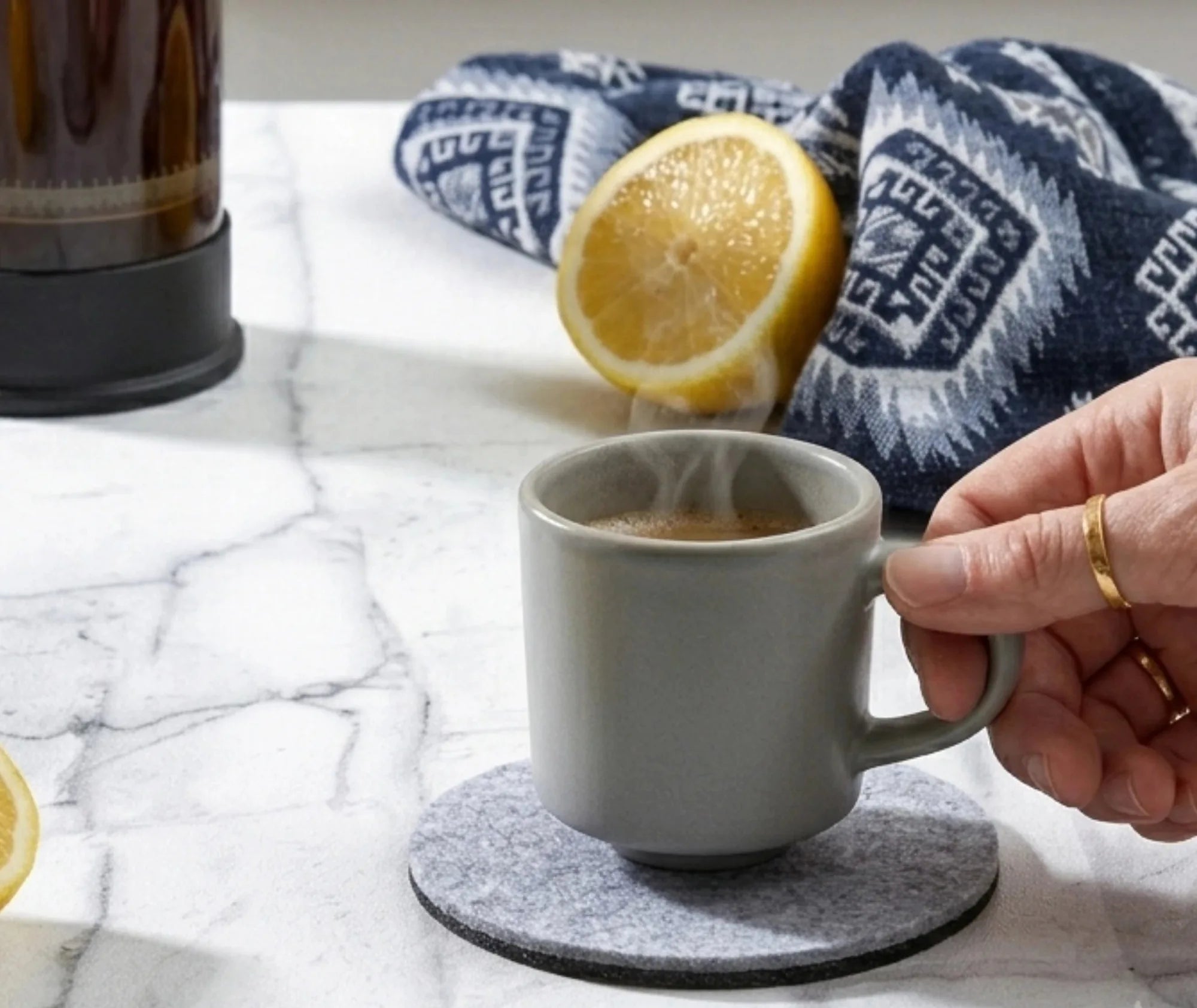 Main soulevant une tasse fumante posée sur un sous-verre en feutrine, avec un citron coupé et un torchon à motifs bleus en arrière-plan.