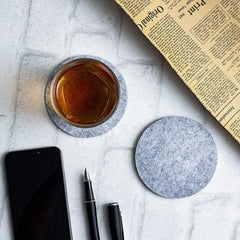 Verre de whisky posé sur un sous-verre en feutrine grise, entouré d'un journal, d'un smartphone et d'un stylo sur une table en marbre.