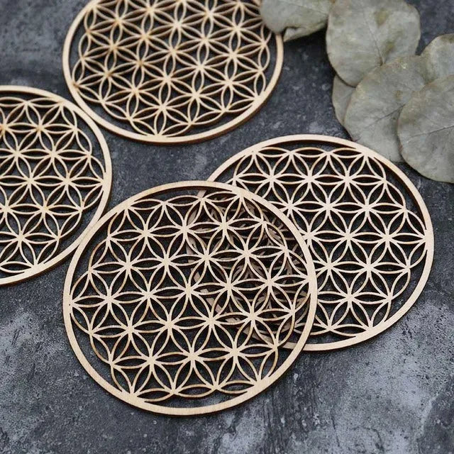 Vue macro de sous-verres en bois motif Fleur de Vie empilés, accompagnés de feuilles vertes décoratives sur fond gris.