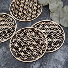 Vue macro de sous-verres en bois motif Fleur de Vie empilés, accompagnés de feuilles vertes décoratives sur fond gris.