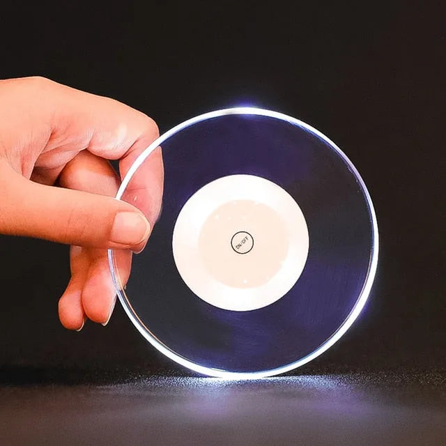 Sous-verre lumineux tenu à la main, disque translucide avec bouton marche/arrêt central et halo lumineux bleuté.