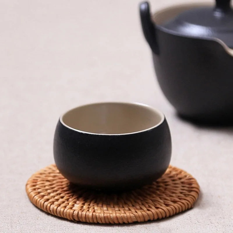 Tasse à thé noire en céramique posée sur un sous-verre en osier tressé, avec une théière noire en arrière-plan.