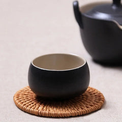 Tasse à thé noire en céramique posée sur un sous-verre en osier tressé, avec une théière noire en arrière-plan.
