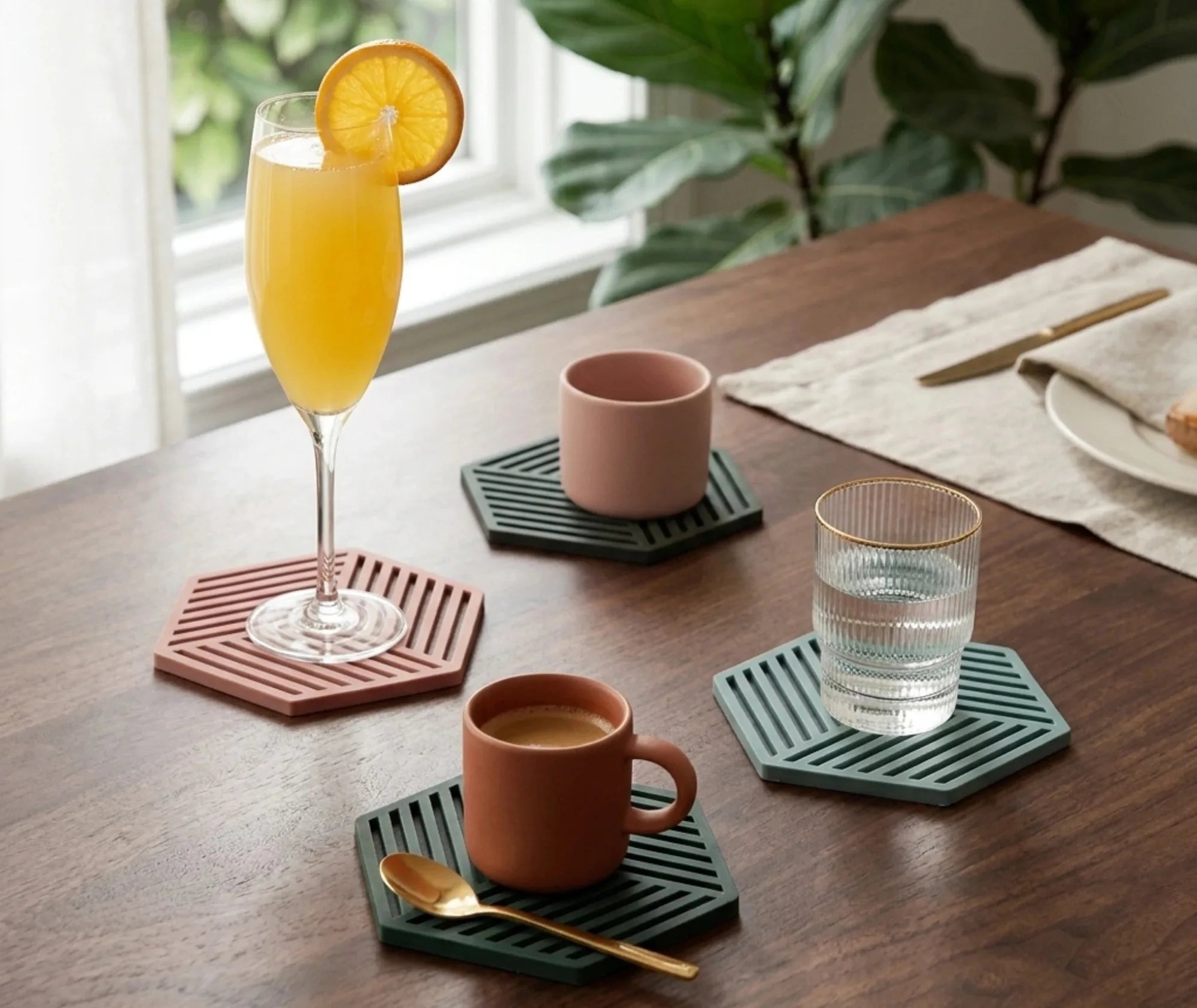Scène de brunch avec une flûte de mimosa, une tasse et un mug en céramique posés sur quatre sous-verres hexagonaux colorés devant une fenêtre.